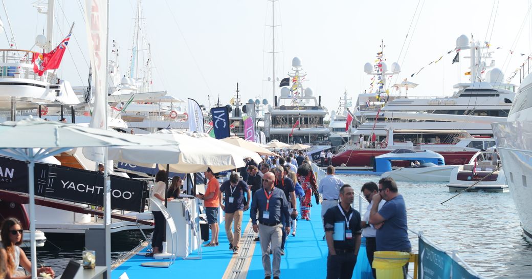 Superyachts on display at Monaco Yacht Show with exhibitor tents along blue carpet boardwalk. Multiple visitors walking between yachts.