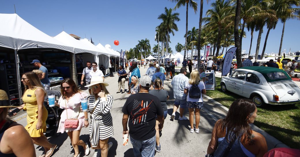 Stands at the Palm Beach International Boat Show with cars lined along right hand side. Many visitors walking along exhibitor tents.