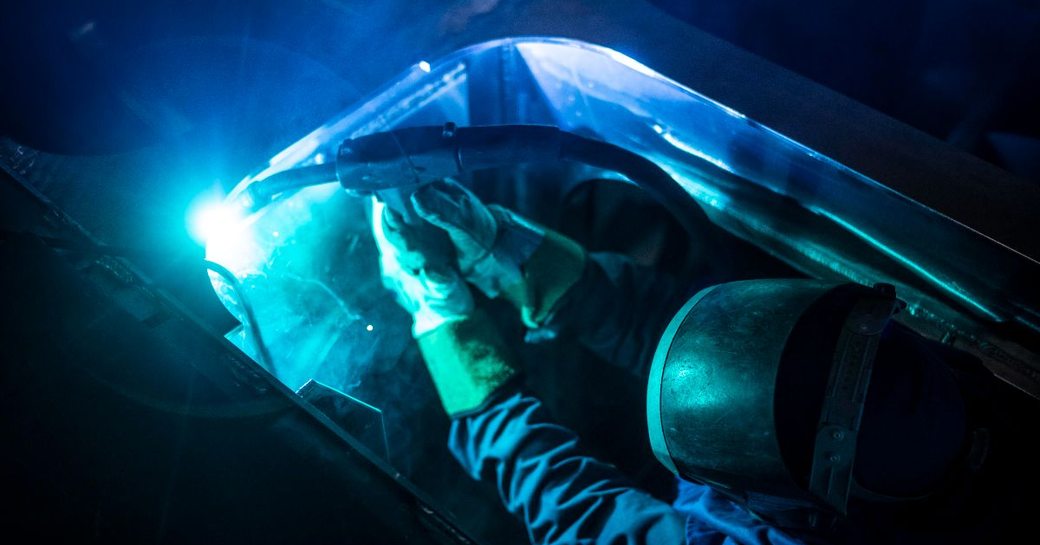 Crew member attaching a keel onto the hull of a yacht. Wearing PPE with electronic tool.