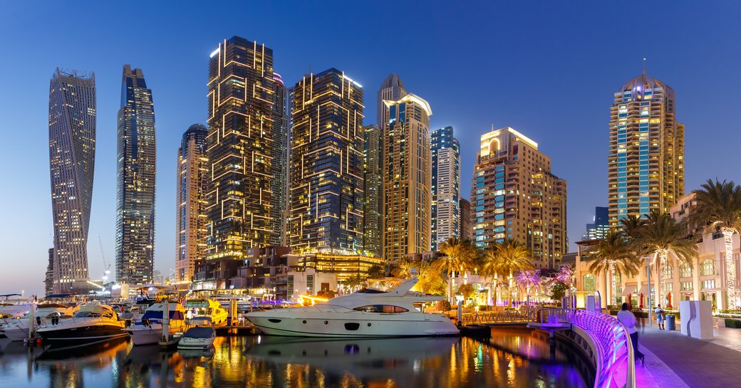 Overview of Dubai Harbour at twilight, motor yachts berthed, with skyscrapers lit up across the sky.