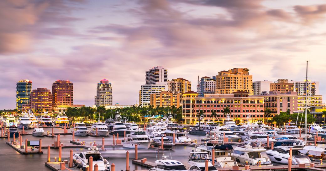 West Palm Beach marina at dusk, with multiple motor yachts berthed. Surrounded by towering hotels and infrastructure.