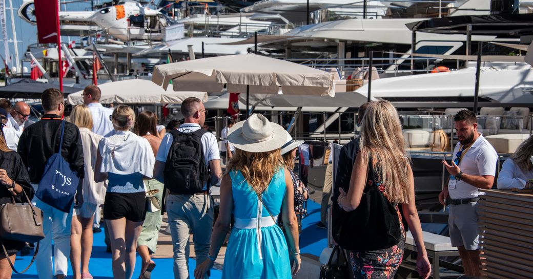 Visitors to the Monaco Yacht Show walking on a blue carpet adjacent to superyachts berthed in Port Hercule