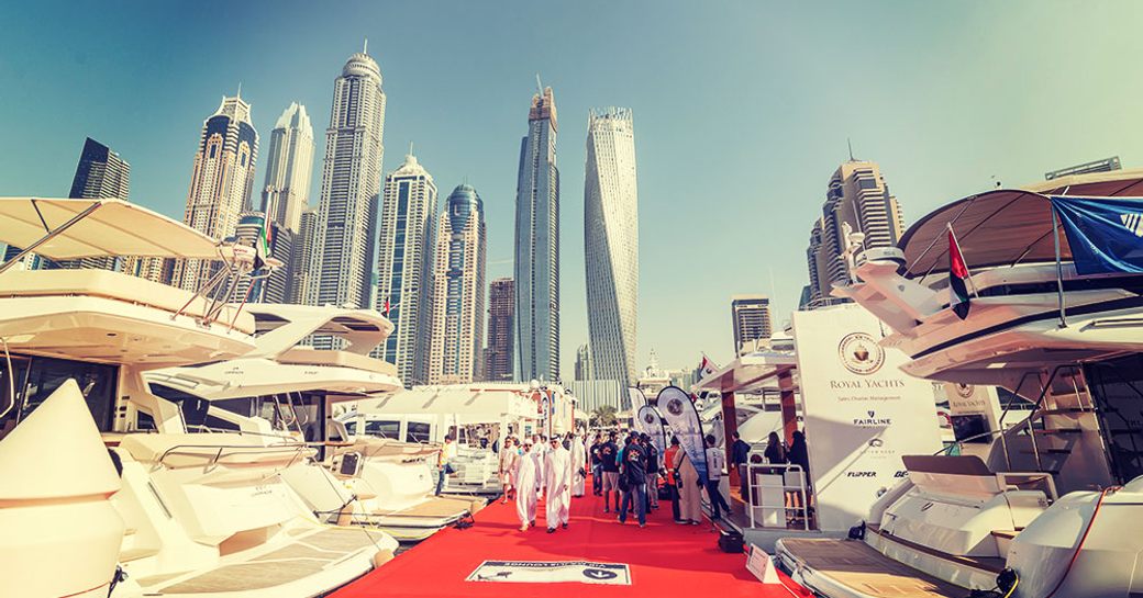 Overview at ground level of Dubai boat show, looking up towards skyscrapers, with yachts berthed either side of red carpeted slip.