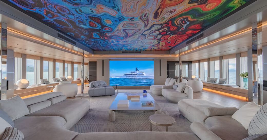 Overview of main salon onboard superyacht WANDERLUST. Multicoloured ceiling design over a cream U-shaped sofa plus extensive glazing to both port and starboard sides.