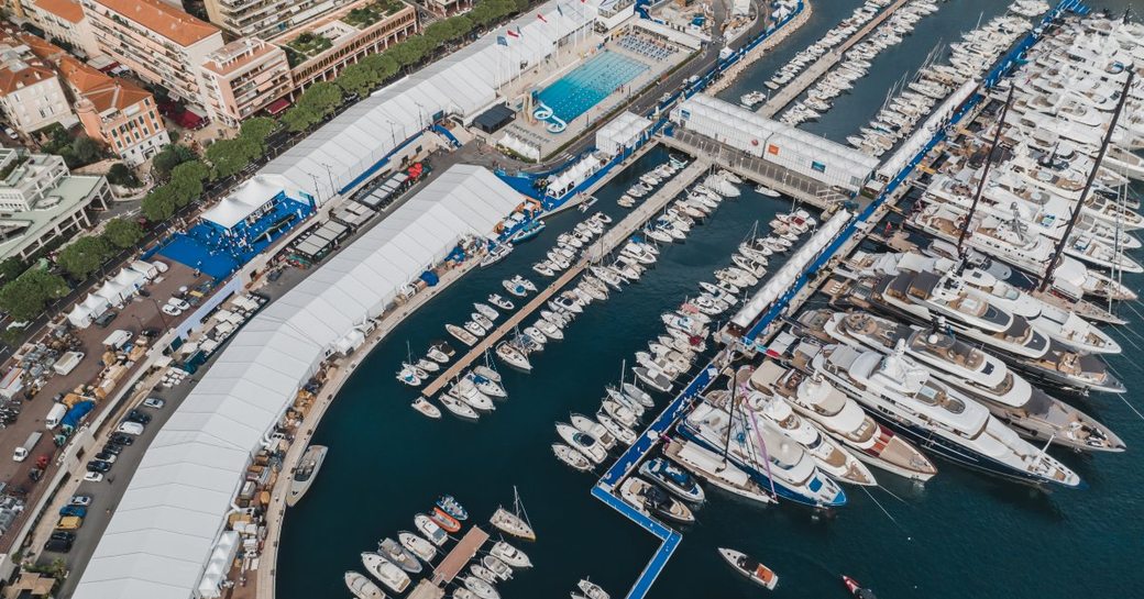 Aerial view of Monaco Yacht Show stands, with superyachts at dock in front and hotels behind.