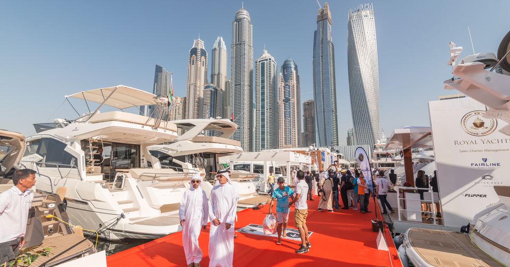 Motoryachts moored on either side of pontoon at Dubai International Boat Show, visitors walking along red carpet with Dubai skyline in background