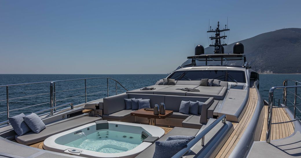 Upper deck relaxation area onboard Pershing 140 'Touch Me', central Jacuzzi surrounded by padded seating and large sun pad, surrounded by sea and elevated terrain to starboard