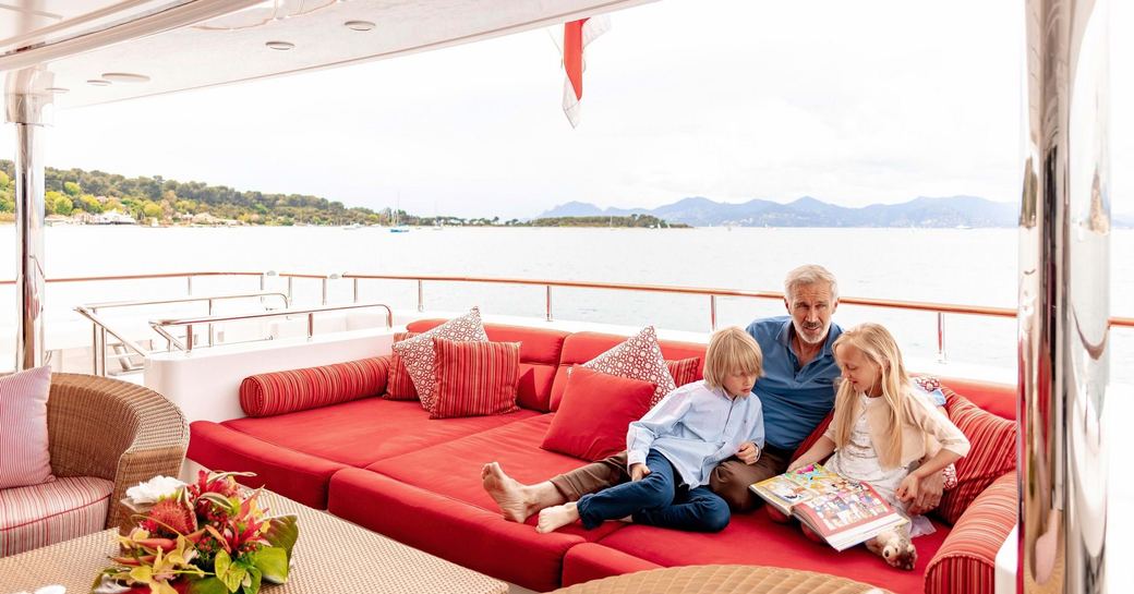 Older man with two young children sitting on large sofa aft of yacht with beautiful landscape behind