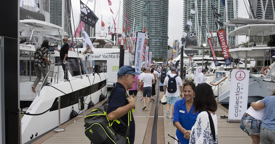 Visitors at the Miami International Boat Show in discussion along the dock with yachts for sale on either side