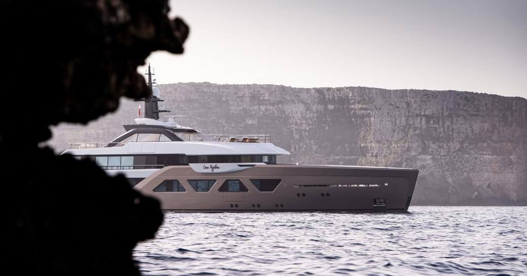 Side view of Amels 60 superyacht Come Together emerging from behind foliage on the sea, with cliffs in the background