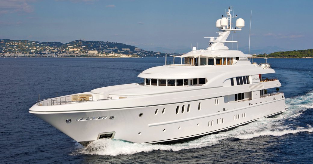 Lurssen yacht CAIPIRINHA for sale at 2024 Monaco Yacht Show and the image shows the yacht making way in a smooth sea on a bright day. The shot is taken forward of the port bow, mid aerial. Land in background. 