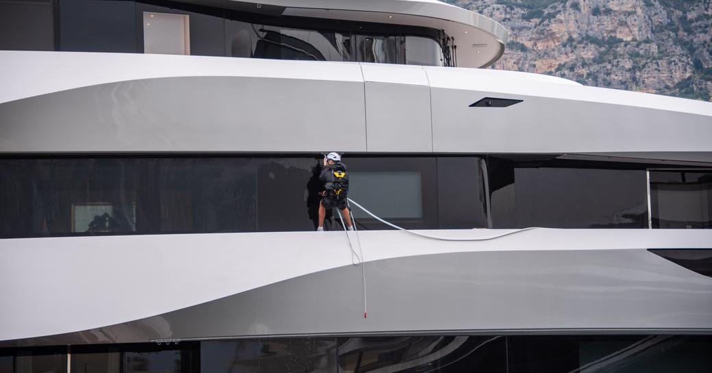 yacht crew preparing a yacht at Monaco Yacht Show, hanging over the side, in a harness and with hard hat, cleaning windows of yacht