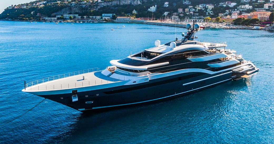 Oceanco yacht DAR shown on anchor in calm sea on bright day with land in background, the shot is mid aerial towards port bow and side profile