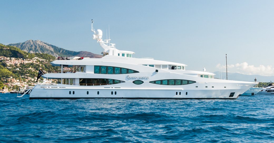 Oceanco yacht QUEEN MAVIA shown on acnhor in calm sea on bright day, shot taken from water level towards starboard side profile, other yachts and land in background