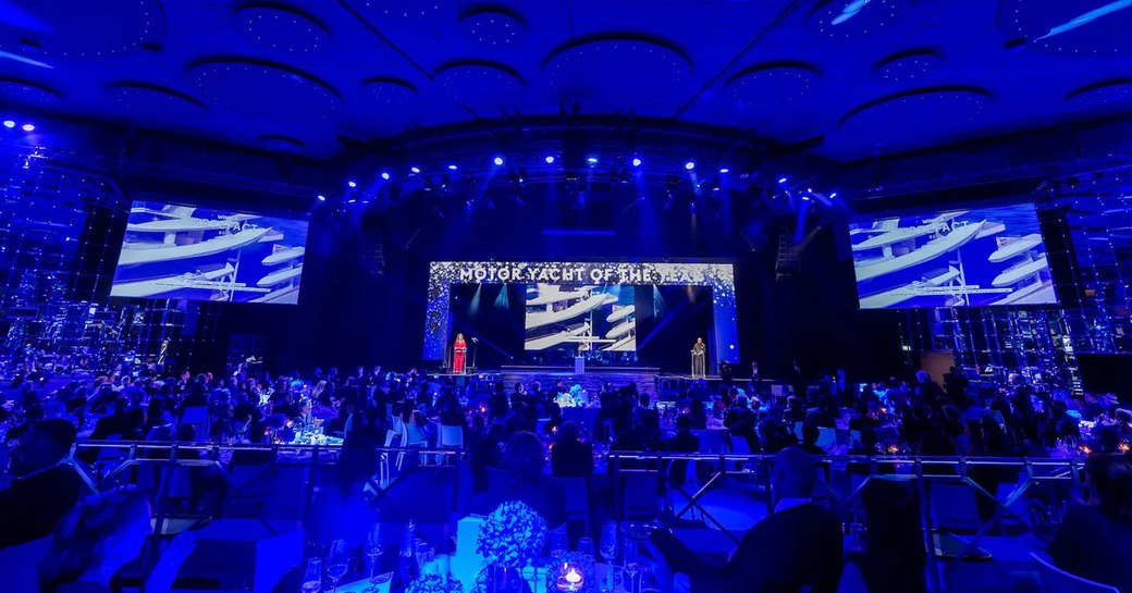 Overview of the World Superyacht Awards ceremony in 2021. Presenter on stage speaking to room full of tables and nominees. 