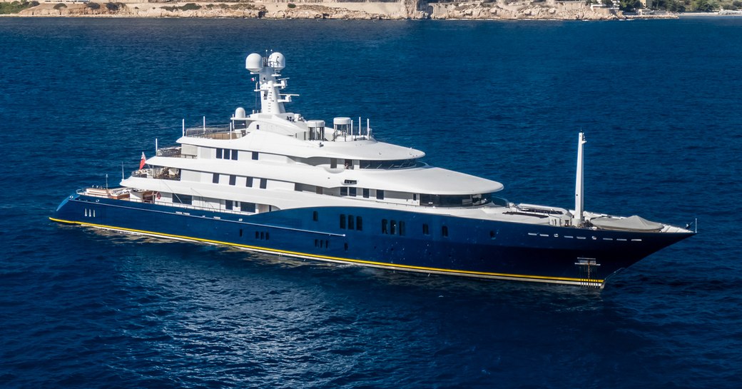 Abeking and Rasmussen superyacht HBC shown in aerial shot on anchor in dark calm waters, bright day, with land in background. The image shows the starboard bow and side profile. 