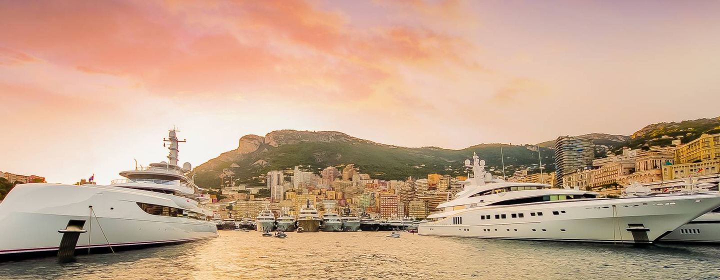 Overview of Port Hercule, Monaco Yacht Show at dusk. Superyachts moored in foreground with Monaco in background.