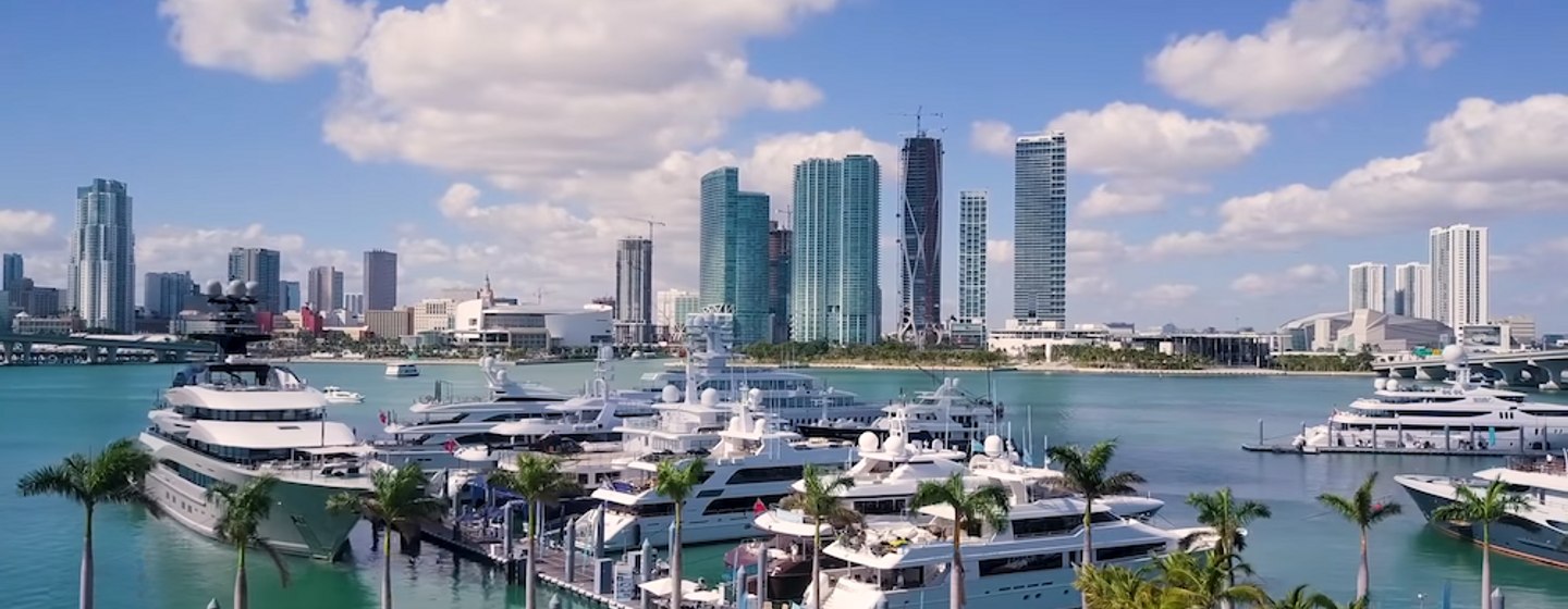 Overview of Yacht Haven Grande Miami at Island Gardens, multiple superyachts moored, looking at skyscrapers over the water.