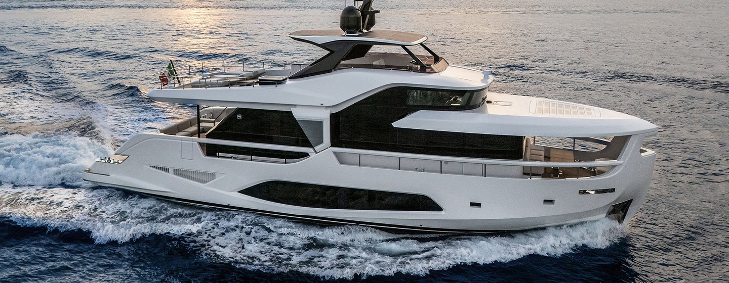 A starboard-side view of the Ferretti Yachts INFYNITO 80 making way through a calm sea at dusk with a rocky landscape in the background