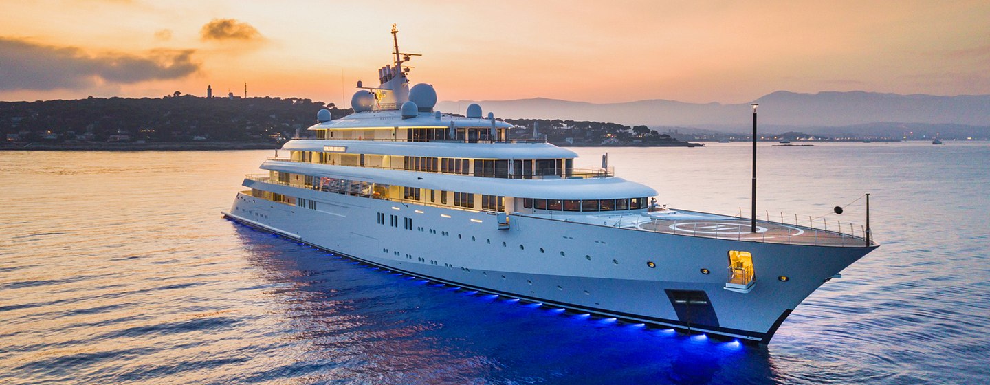 Lurssen yacht GOLDEN ODYSSEY shown at sunrise, anchored in calm waters with land in background, underwater waters show the perimeter of the yacht in the water