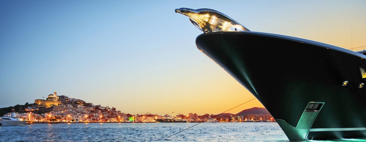 PHOENIX 2 Lurssen yacht bow with iconic figurehead of phoenix, shot taken from water, looking up a bow at sunset, illuminated land in background