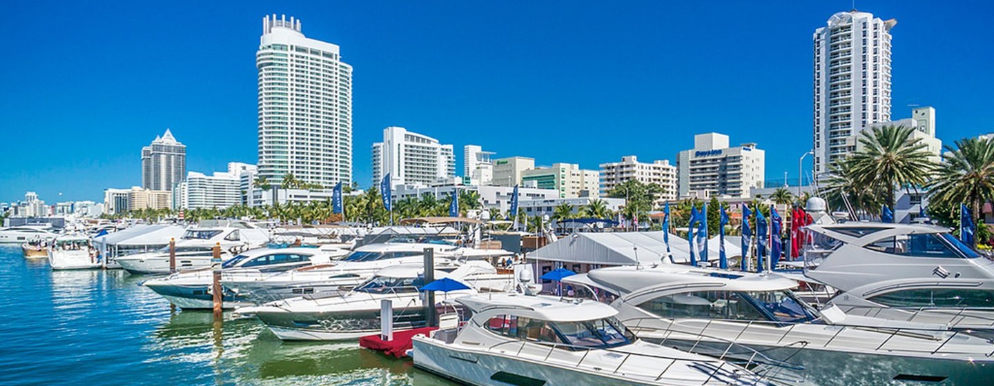 Water level view of Miami International Boat Show, yachts moored in marina with skyscrapers in background.