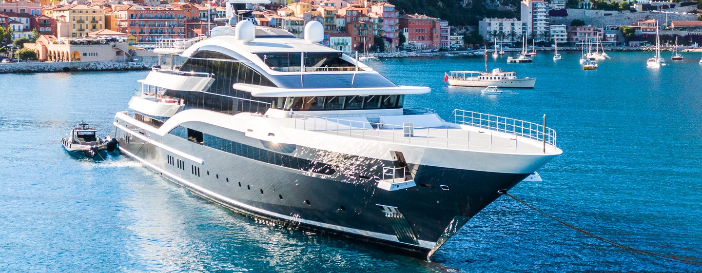 Oceanco yacht DAR shown at anchor in blue sea on bright day, tender off the starboard aft deck, land in background