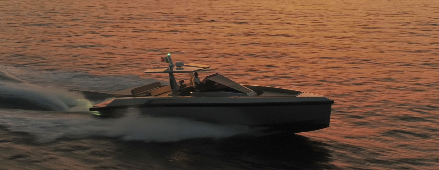 Tender boat moving fast on water at dusk with coastline in the background.