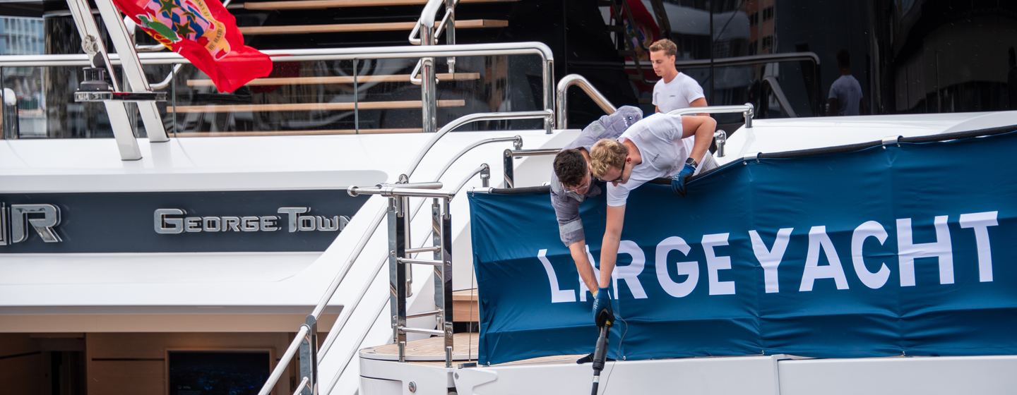 yacht crew at monaco yacht show 2024 preparing a yacht for show, there is a big sign saying LARGE YACHT that the crew are fastening to the back of the yacht