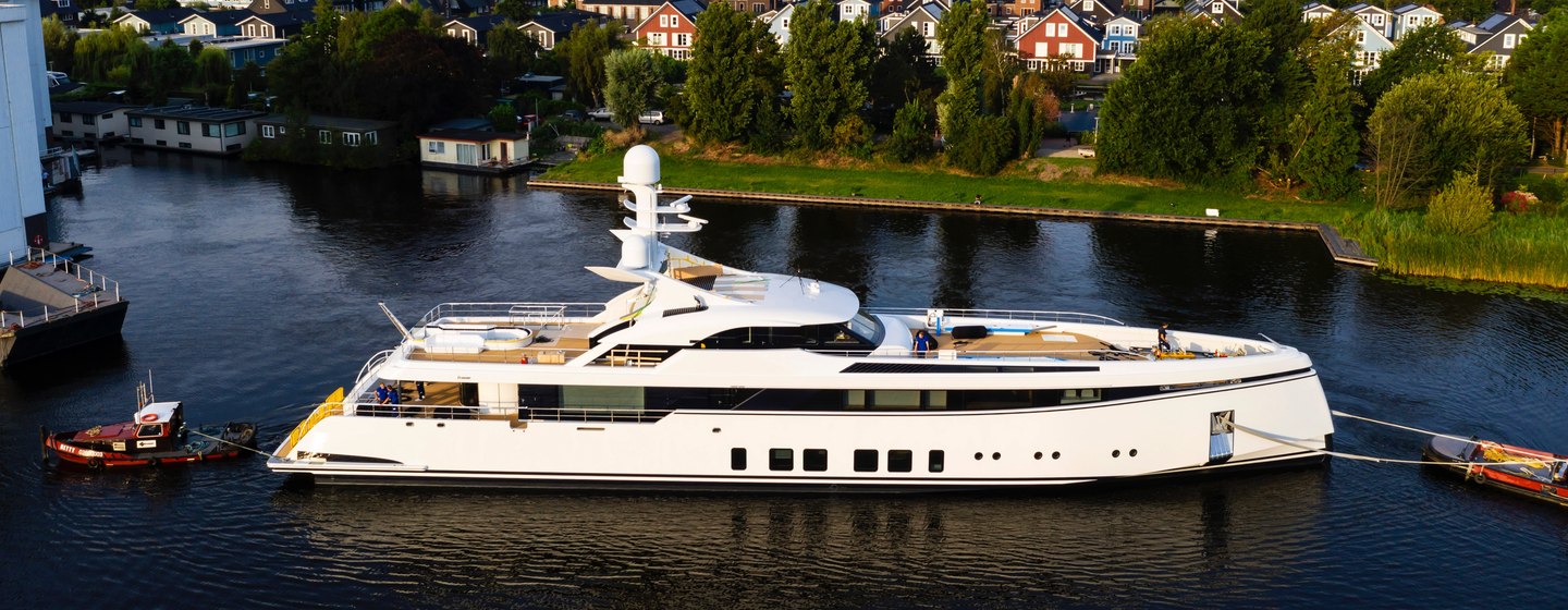 Feadship Project 706 being escorted out of construction yard, surrounded by foliage and river