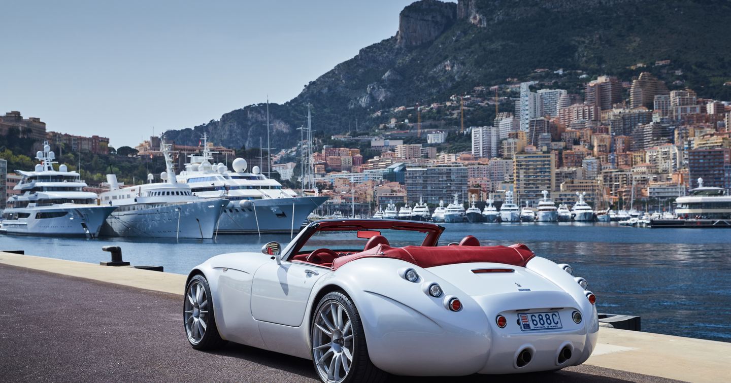 Motor car on promenade alongside Port Hercule. Multiple motor yachts berthed in marina with Monte Carlo in background.