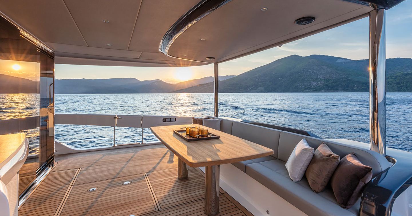 Aft deck onboard Navetta 64, seating along rear side with table. Surrounded by sea and elevated terrain in distance.