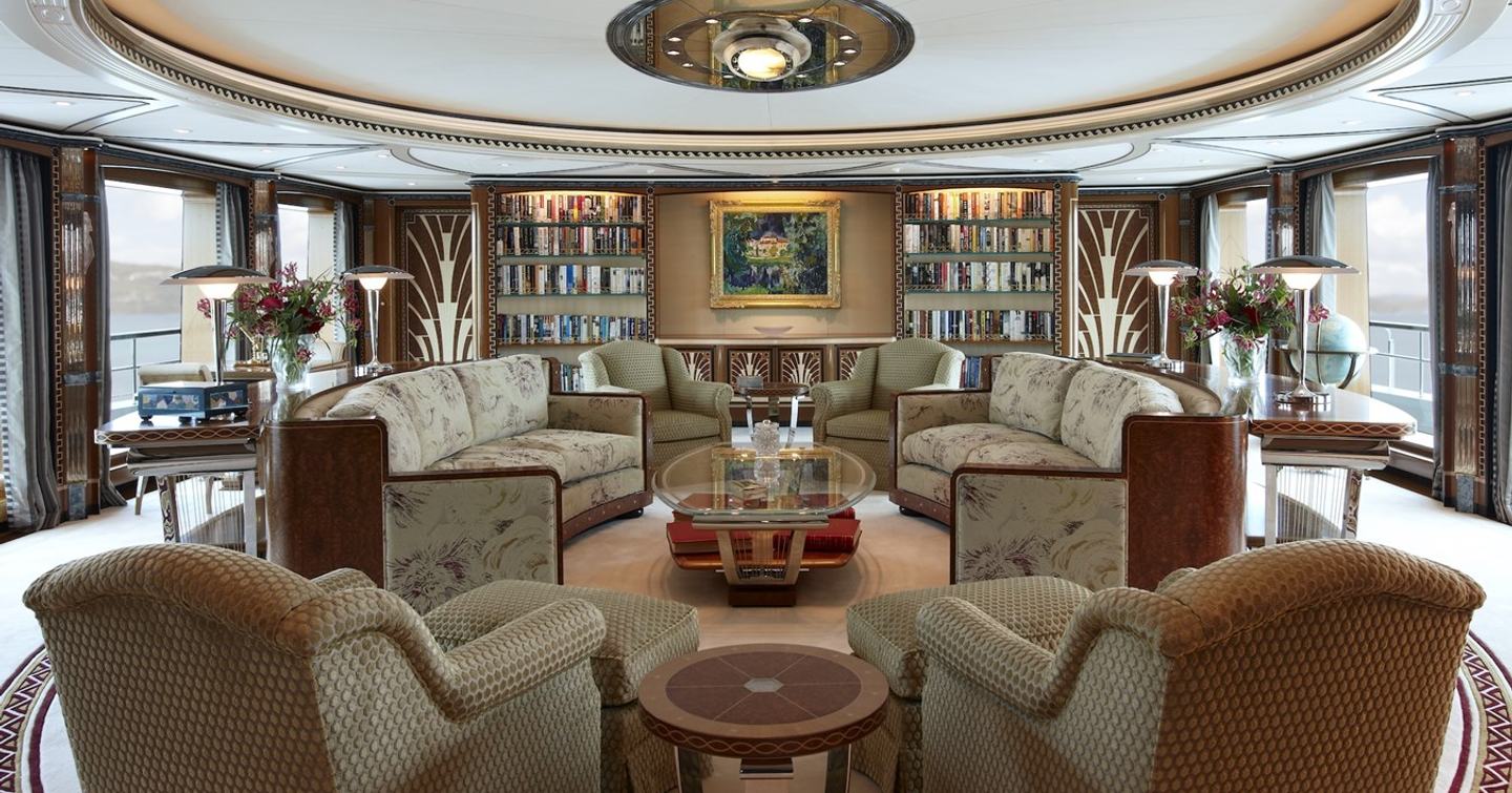 Elegant main saloon aboard Lady Christine featuring Art Deco design, plush seating, circular layout, bookshelves, and panoramic windows with sea views.