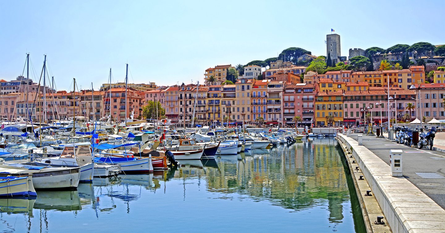 Water level view across Cannes marina, overlooking the Old Town. Sailboats moored to port side with walkway on starboard.