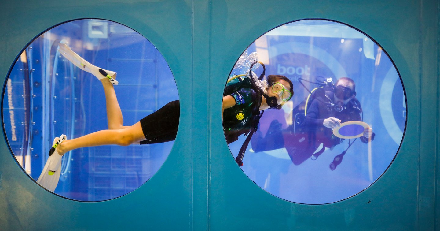 Diving demonstration at boot Dusseldorf show. Female diver visible in large tank through two adjacent circular windows.