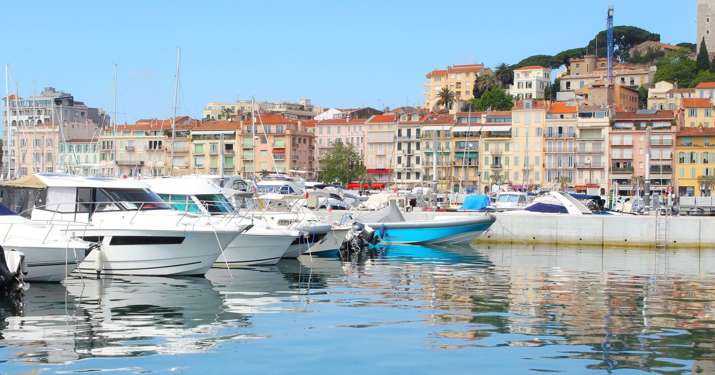 Water level view of Vieux Port in Cannes, several small motor yachts moored to port side, overview of Cannes in background.