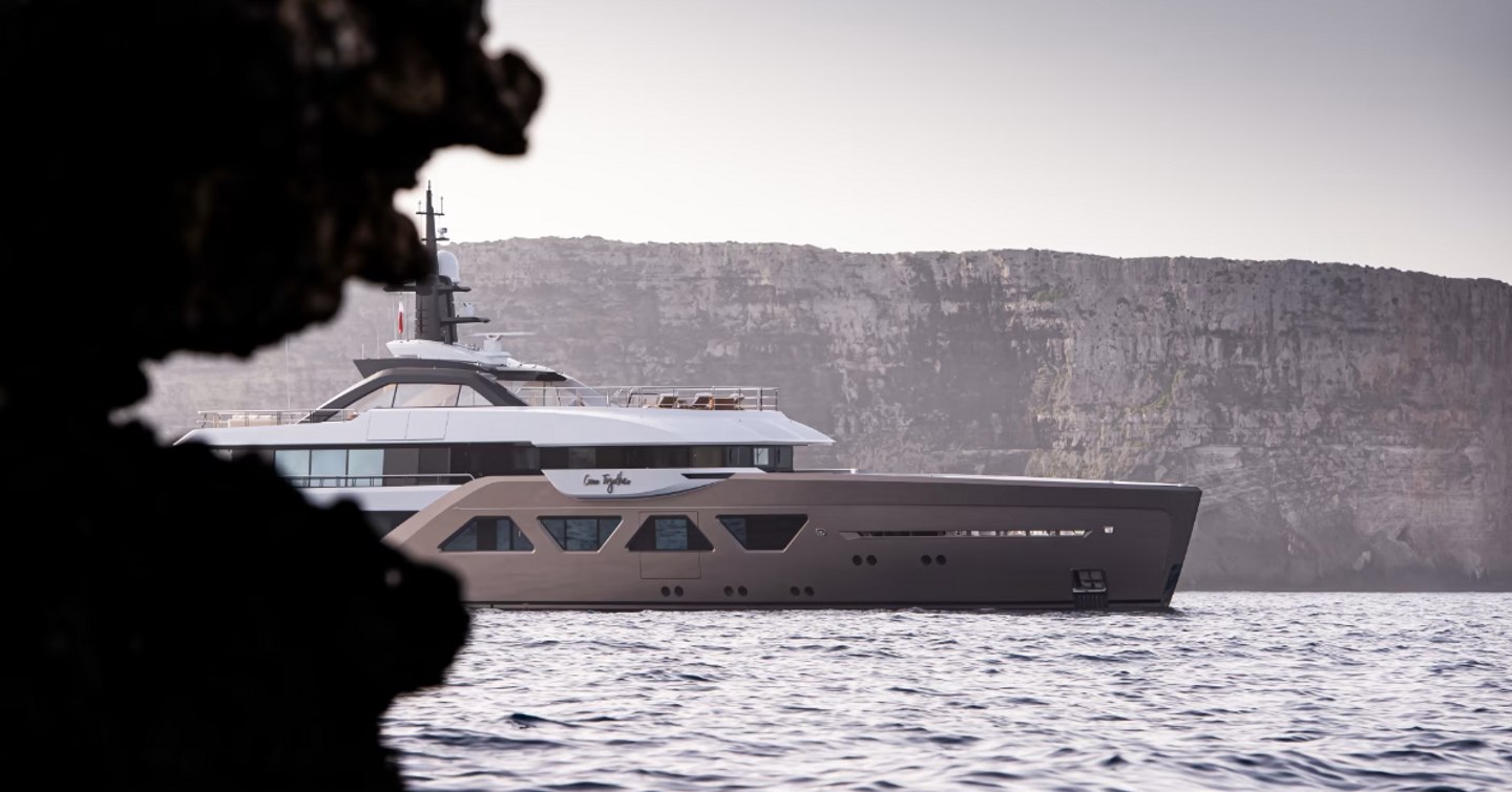 Side view of Amels 60 superyacht Come Together emerging from behind foliage on the sea, with cliffs in the background