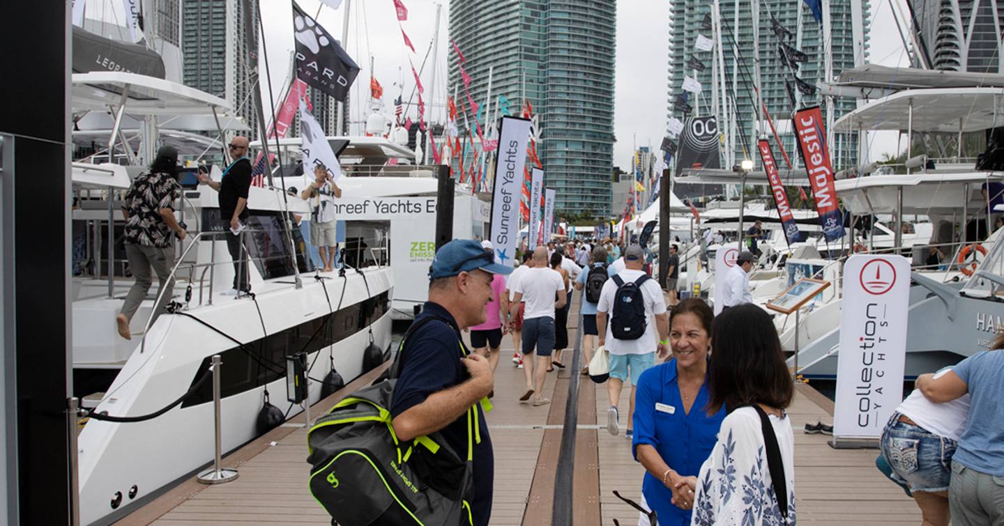 Visitors at the Miami International Boat Show in discussion along the dock with yachts for sale on either side