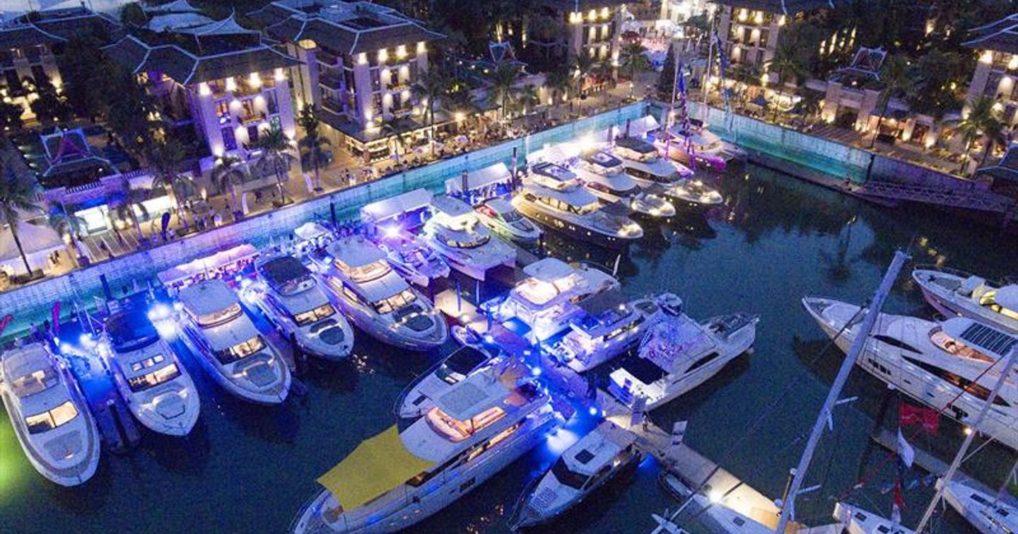 Motor yachts berthed at Thailand Yacht Show at dusk, neon blue lights illuminate water.