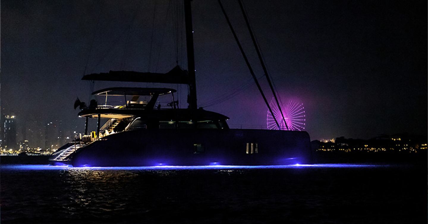 Silhouette of a Sunreef Yacht catamaran at night. Blue lights illuminate the sea line with a cityscape in the background.