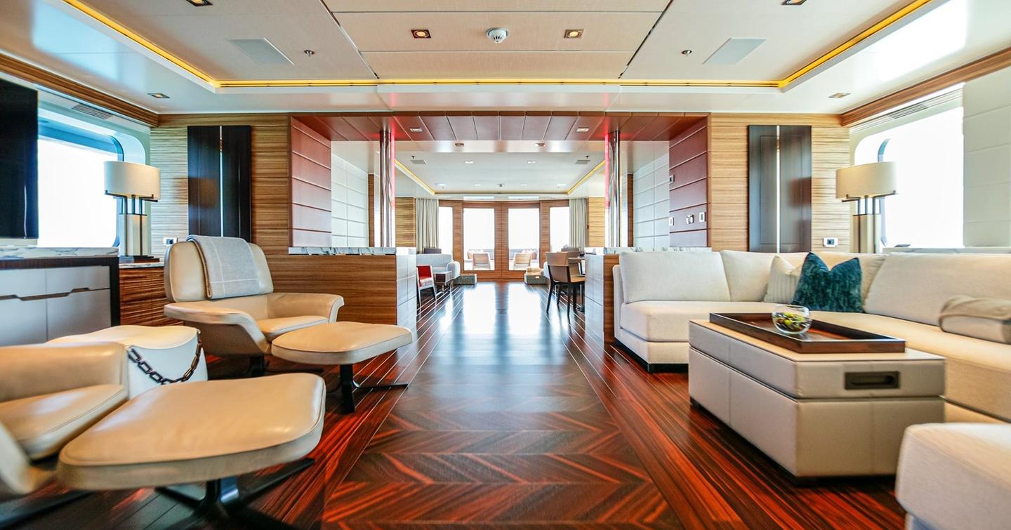Motor yacht W's main saloon with polished wood flooring, cream sofas, armchairs, and large windows bringing in natural light.