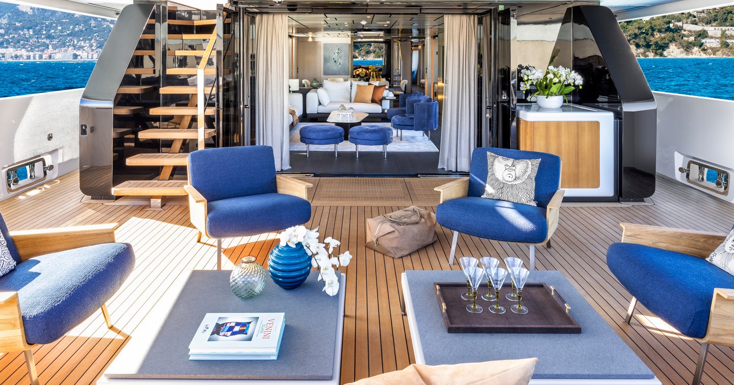 Aft deck onboard Custom Line 140. Low coffee tables surrounded by blue seating, interior visible through open doors.