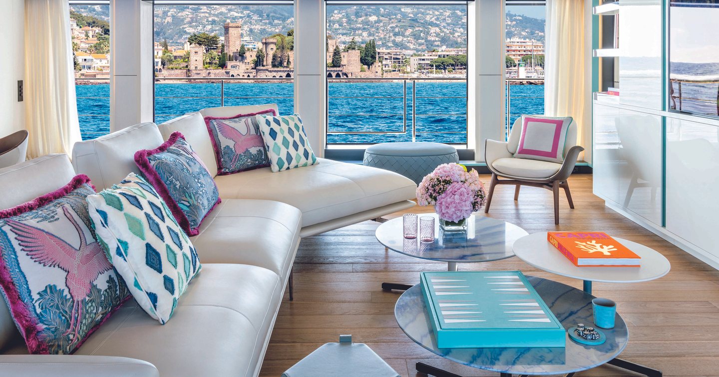 Main salon onboard CRN RIO superyacht, white sofas and vibrant cushions face flatscreen TV, full length windows in background.