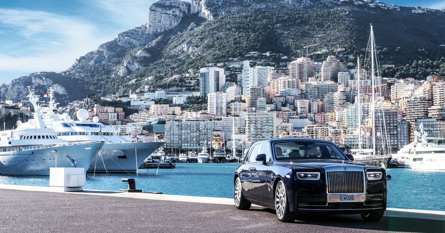 Prestige car parked on dock in front of Port Hercule, superyachts berthed in background.