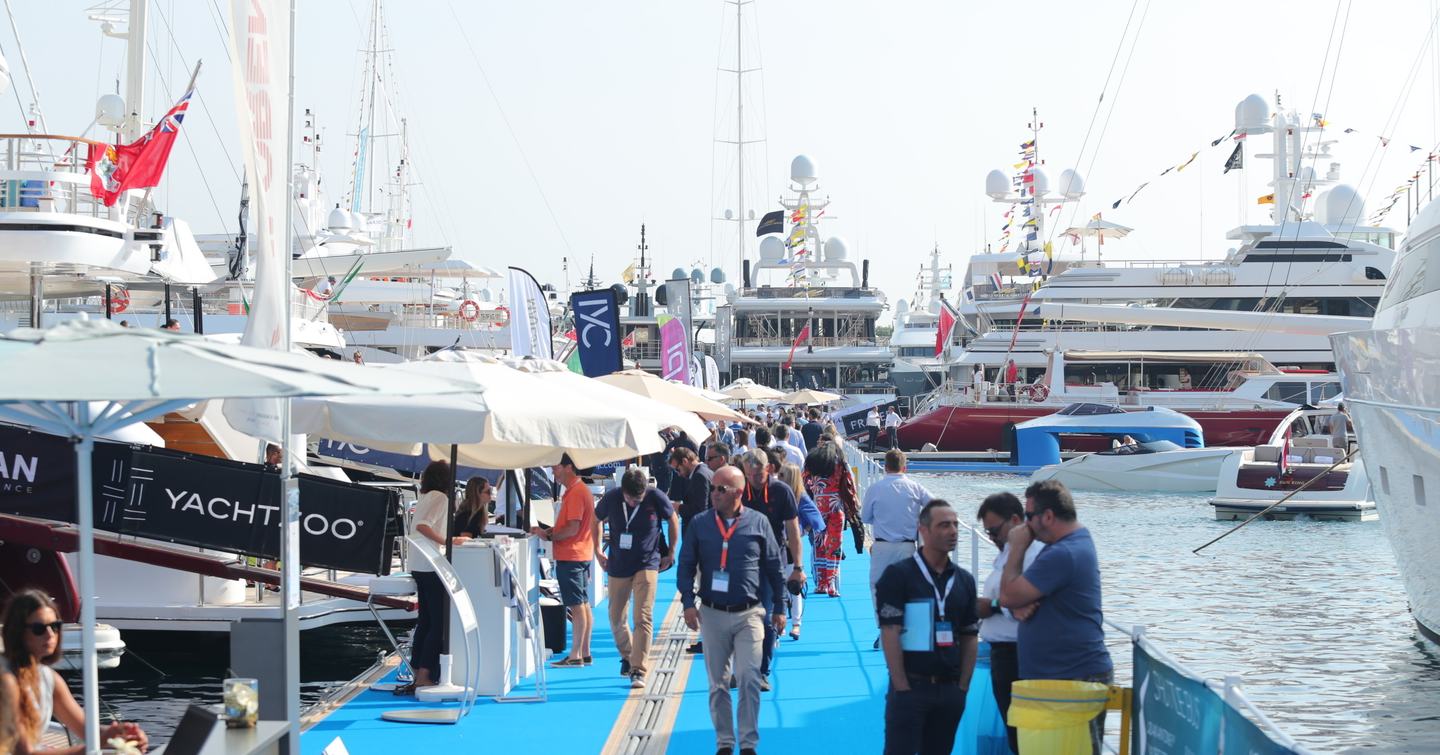 Superyachts on display at Monaco Yacht Show with exhibitor tents along blue carpet boardwalk. Multiple visitors walking between yachts.