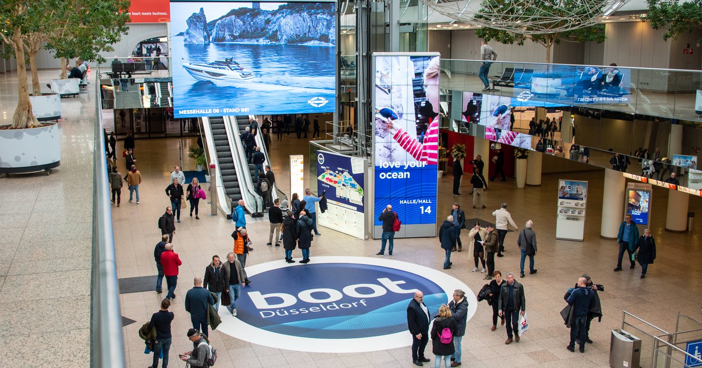 Inside of Messe Dusseldorf during Boot Dusseldorf event. Large logo mat in centre of walkway with lots of visitors milling around into hall entrances.