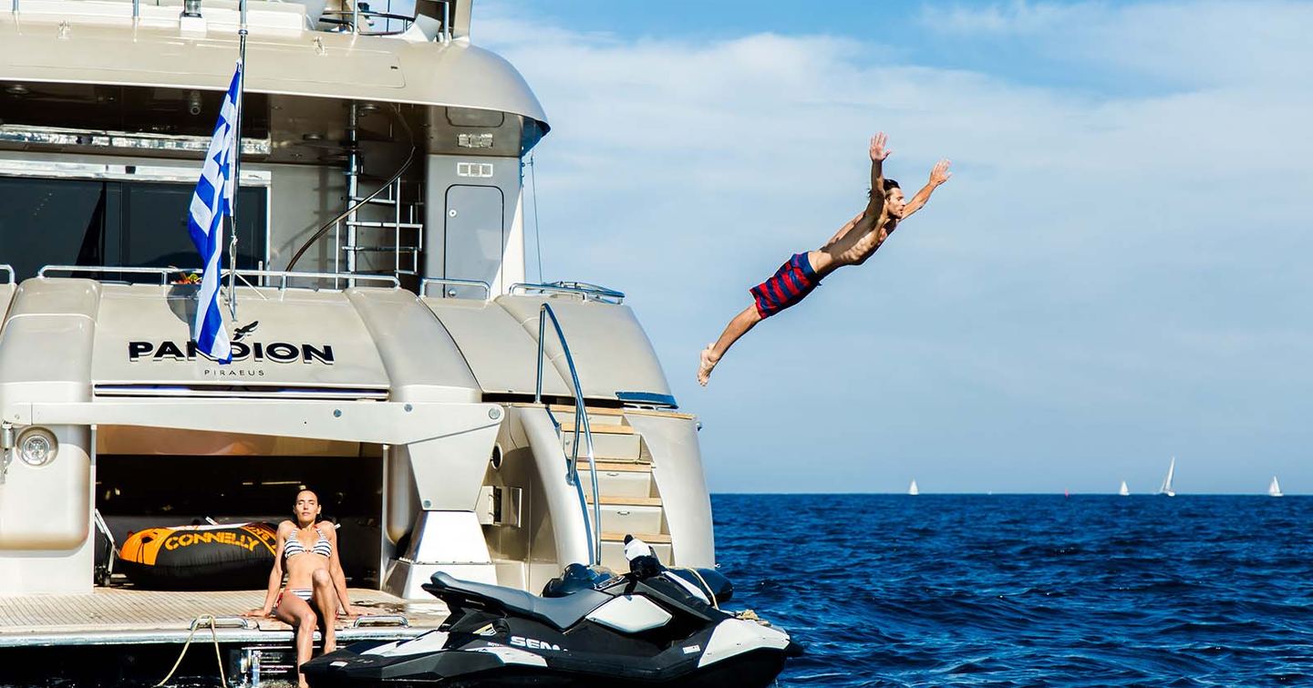 Young female sat on swim platform on yacht with young man in mid-air arms outstretched diving into water. PWC is visible in front of the yacht