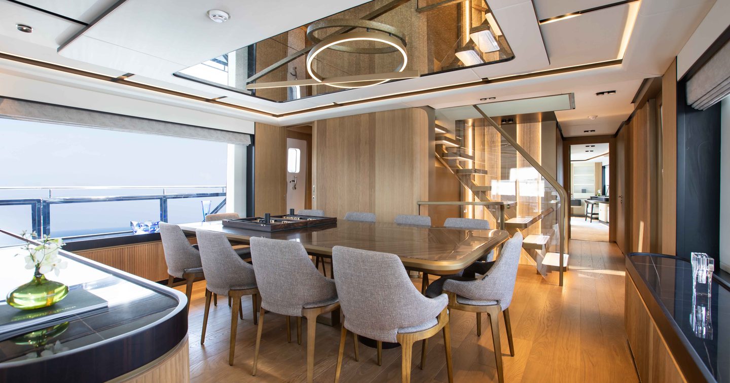 Dining area onboard the Majesty 111, long table surrounded by white chairs, with a wide window adjacent.