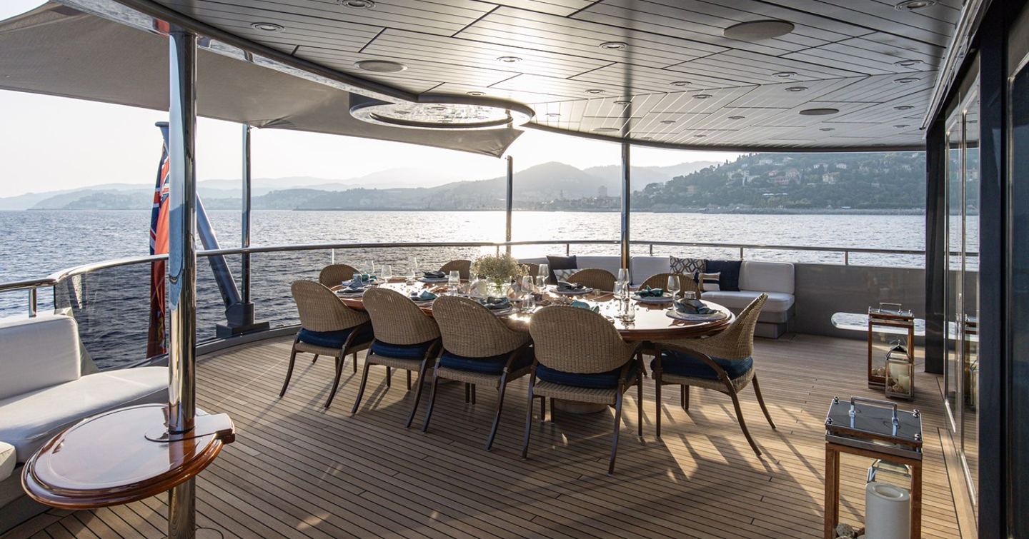 Motor yacht Vibrance's aft deck dining area with set table, wicker chairs, and sea view at sunset.