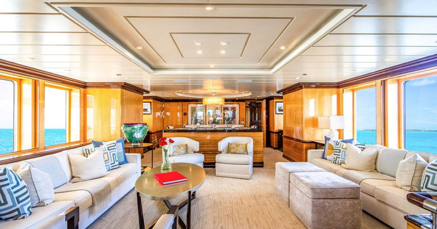 Main saloon on motor yacht Nita K II with large windows, cream sofas, patterned cushions, central coffee table, and a bar at the far end